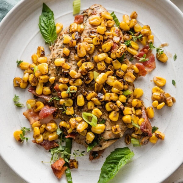 Plate with a helping of chicken breast pan-fried and topped with brown butter corn, bacon, and fresh herbs and green onions.