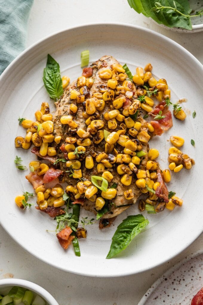 Plate with a helping of chicken breast pan-fried and topped with brown butter corn, bacon, and fresh herbs and green onions.