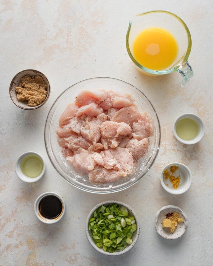 Overhead image of chicken pieces tossed with salt, pepper, and cornstarch; orange juice, brown sugar, ginger, garlic, orange zest, soy sauce, lemon juice, and green onions.