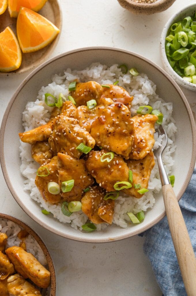 Bowl of easy homemade Asian-inspired orange chicken with a sweet and sticky sauce, served over rice and with green onions and sesame seeds.