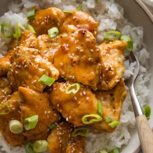 Close up of a bowl of homemade Asian-inspired orange chicken topped with green onions and sesame seeds.