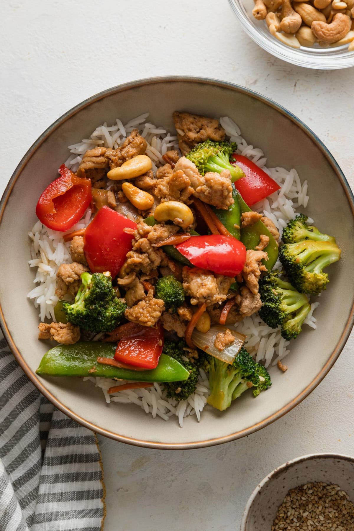 Countertop set with a bowl of ground chicken stir fry served over rice, with cashews and sesame seeds available in small bowls on the side for an optional sprinkle on top.