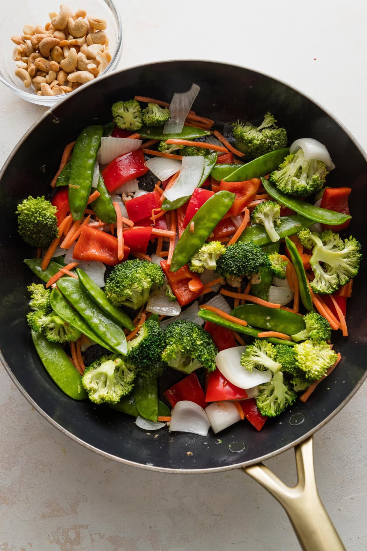 Broccoli florets, bell pepper, shredded carrots, and snow peas added to chopped onion in a pan.