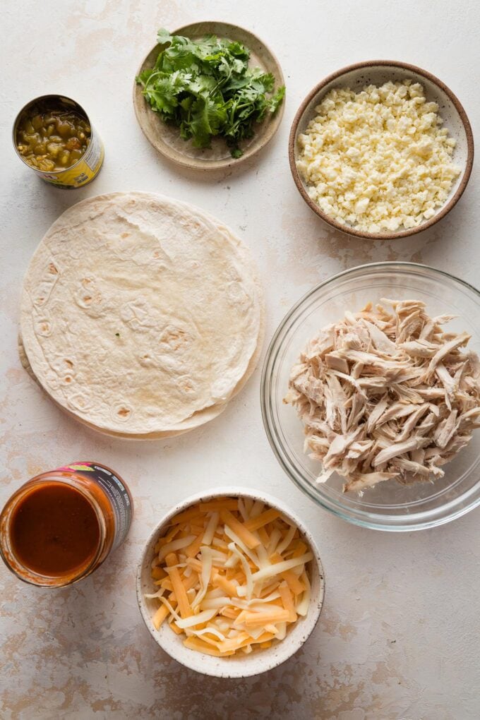 Overhead view of tortillas, shredded chicken, shredded Mexican cheese, crumbled Cotija, enchilada sauce, green chiles, and cilantro leaves.