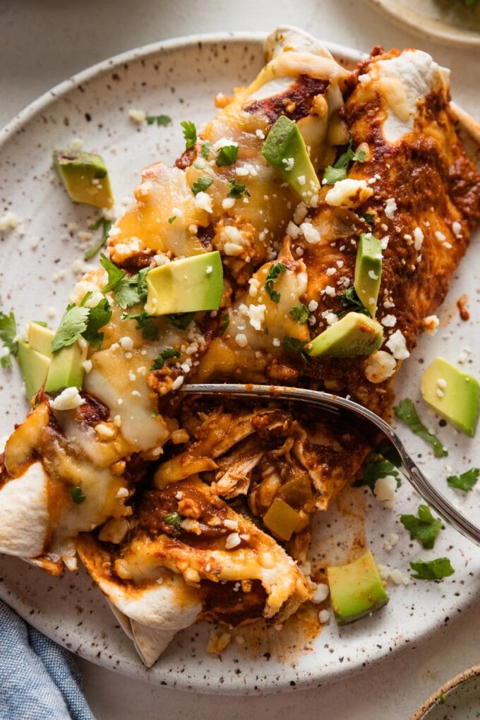 Fork pulling off a bite of rotisserie chicken enchilada from a small dinner plate to show the sauce-coated shredded chicken inside.