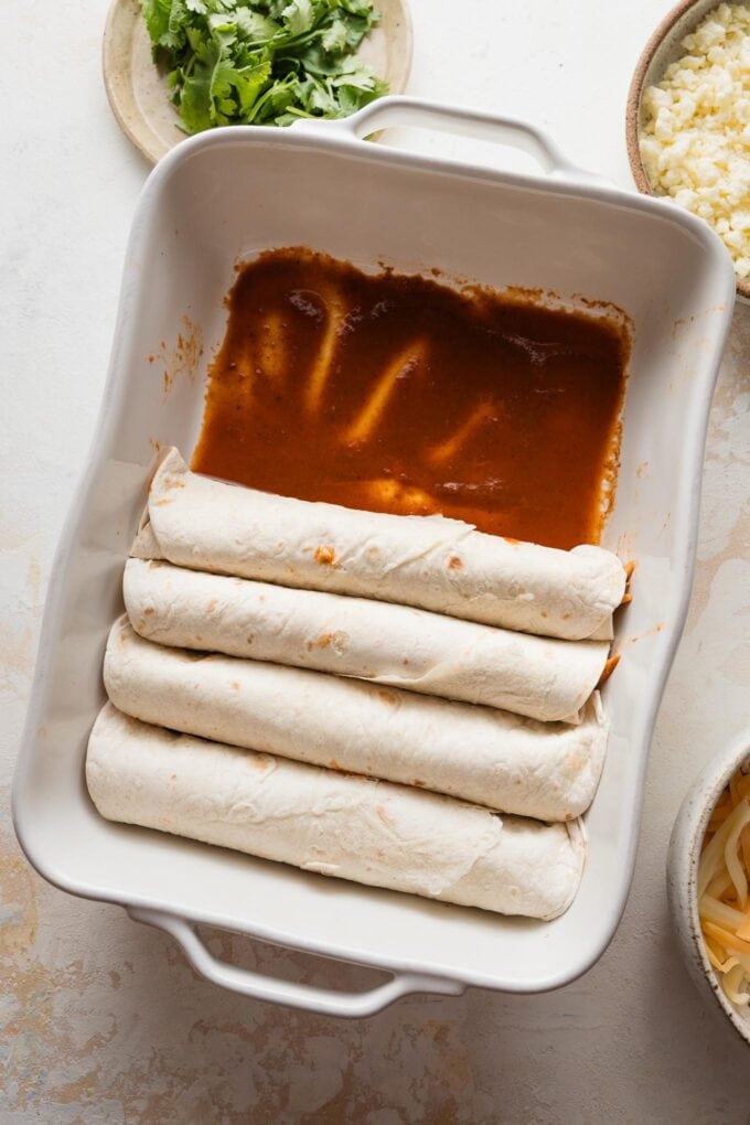 Baking tray partially full of rolled enchiladas tucked in snugly over a thin layer of enchilada sauce.