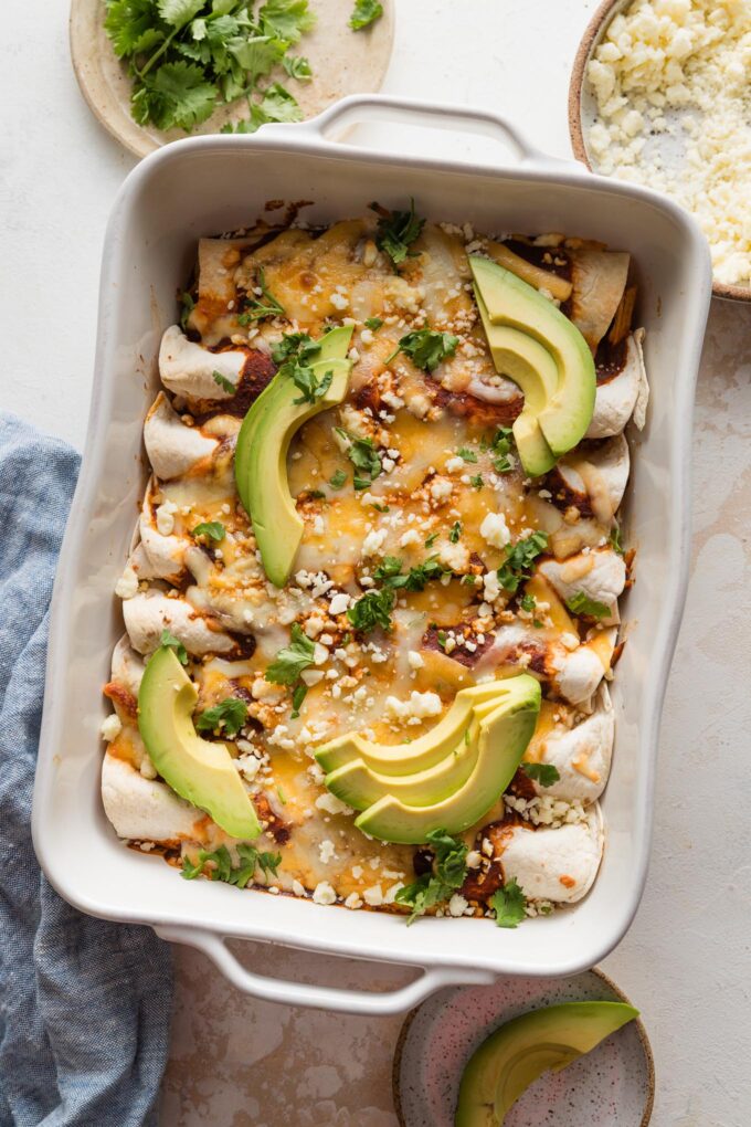 White ceramic pan full of easy rotisserie chicken enchiladas topped with Cotija, cilantro, and avocado.