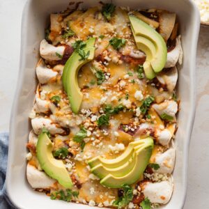 Large ceramic baking dish filled with quick rotisserie chicken enchiladas topped with avocado.