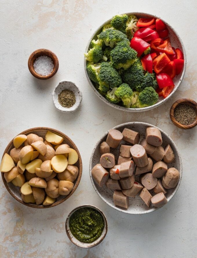 Overhead view of sliced, pre-cooked chicken sausage links, chopped broccoli florets and bell peppers, baby potatoes quartered into small pieces, fresh pesto, and salt and pepper.