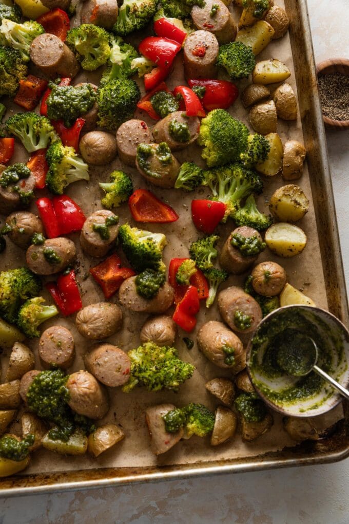 Large rimmed sheet pan lined with parchment and filled with a one pan meal of chicken sausage, potatoes, lots of veggies, and pesto.