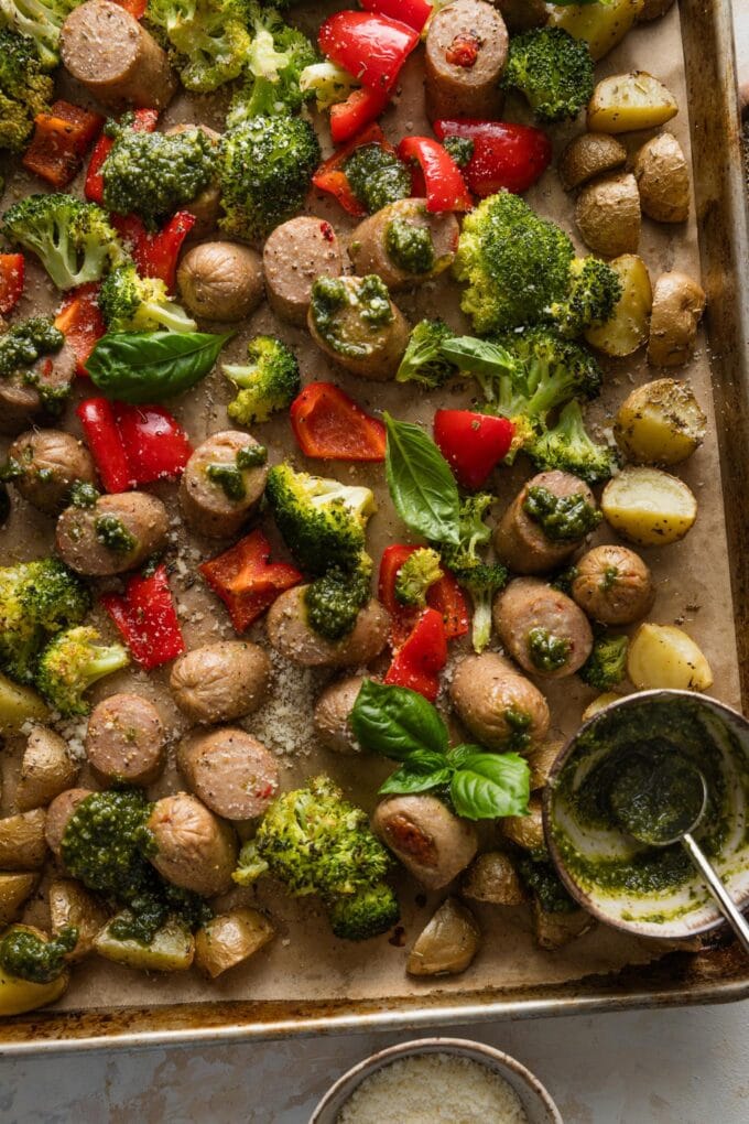 Large rimmed sheet pan lined with parchment and filled with a one pan meal of chicken sausage, potatoes, lots of veggies, and fresh basil pesto resting in a small bowl on the pan and dolloped over everything else.
