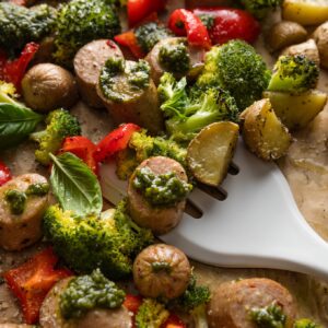Spatula lifting a portion of a one sheet pan meal with chicken sausage, broccoli, peppers, potatoes, and big dollops of fresh basil pesto.