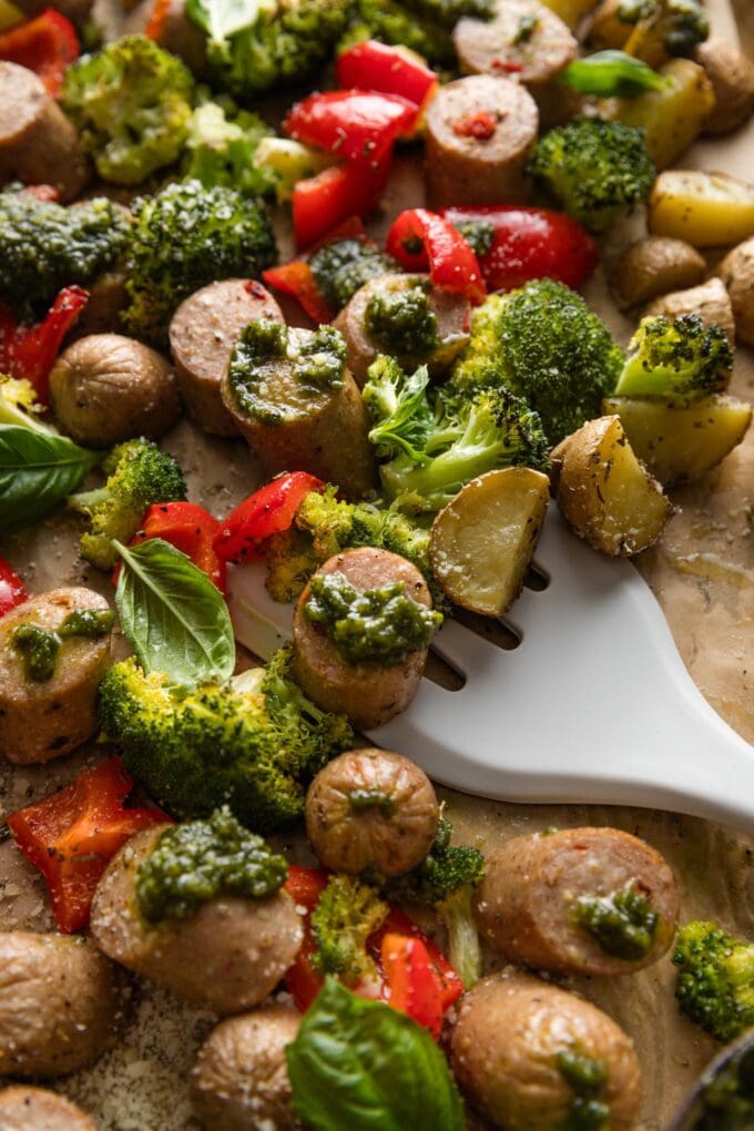 Spatula lifting a portion of a one sheet pan meal with chicken sausage, broccoli, peppers, potatoes, and big dollops of fresh basil pesto.