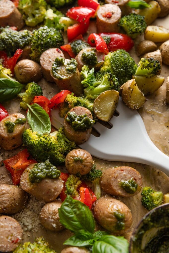 Spatula lifting a portion of a one sheet pan meal with chicken sausage, broccoli, peppers, potatoes, and big dollops of fresh basil pesto.