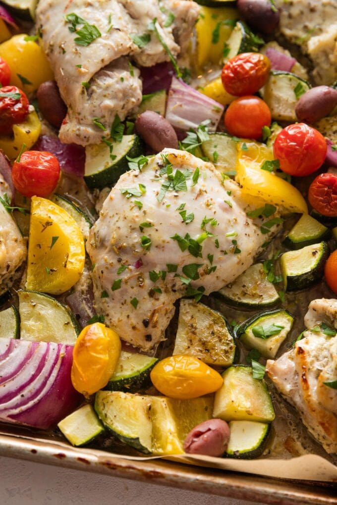 Close up of a chicken thigh with Greek seasonings roasted on a sheet pan with veggies.