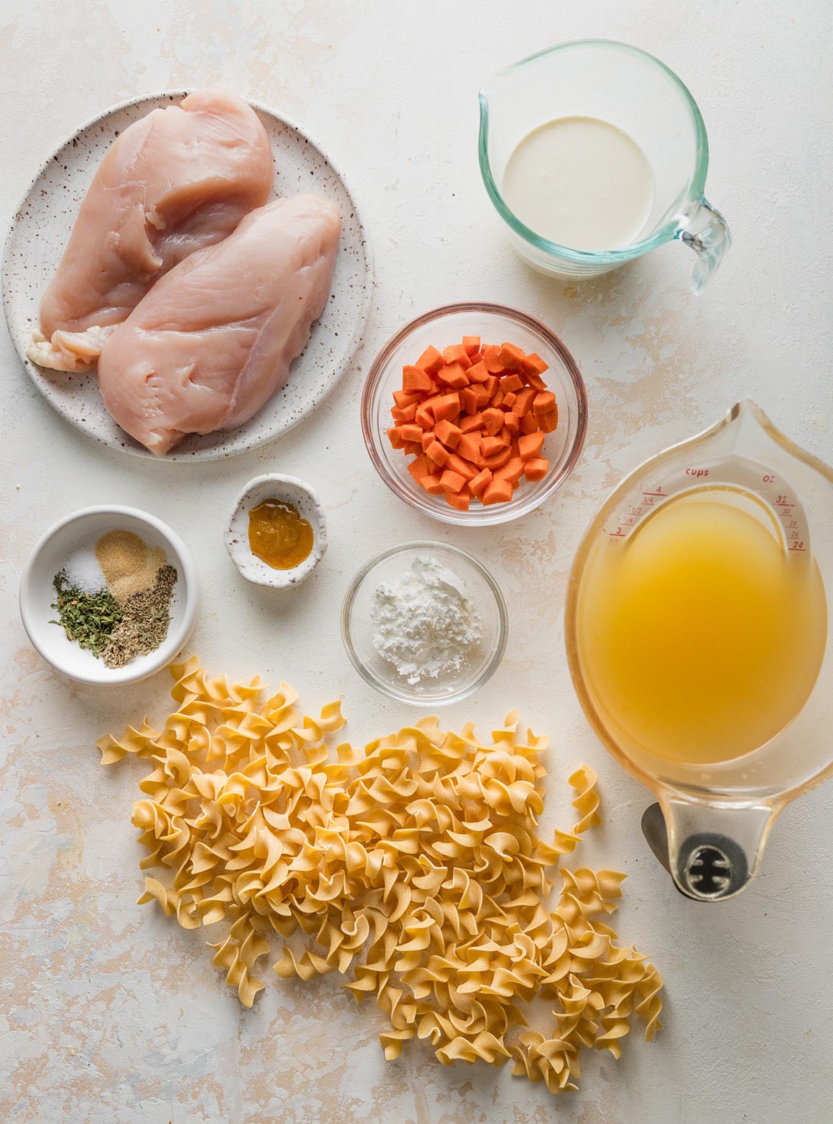 Overhead view of large chicken breasts, chopped carrots, broth, bouillon paste, cream, spices, and egg noodles, all measured into prep bowls and ready to cook.