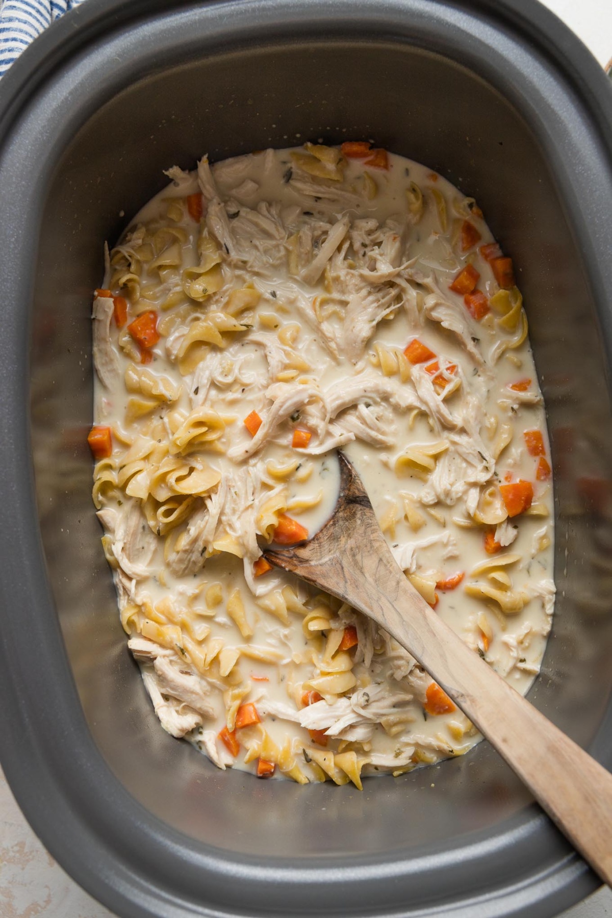 A large slow cooker full of a one pot meal of shredded chicken and egg noodles in a cream sauce, with a rustic wooden spoon dipped in to stir and start serving.