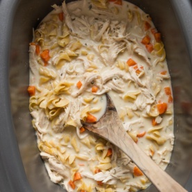 A large slow cooker full of a one pot meal of shredded chicken and egg noodles in a cream sauce, with a rustic wooden spoon dipped in to stir and start serving.