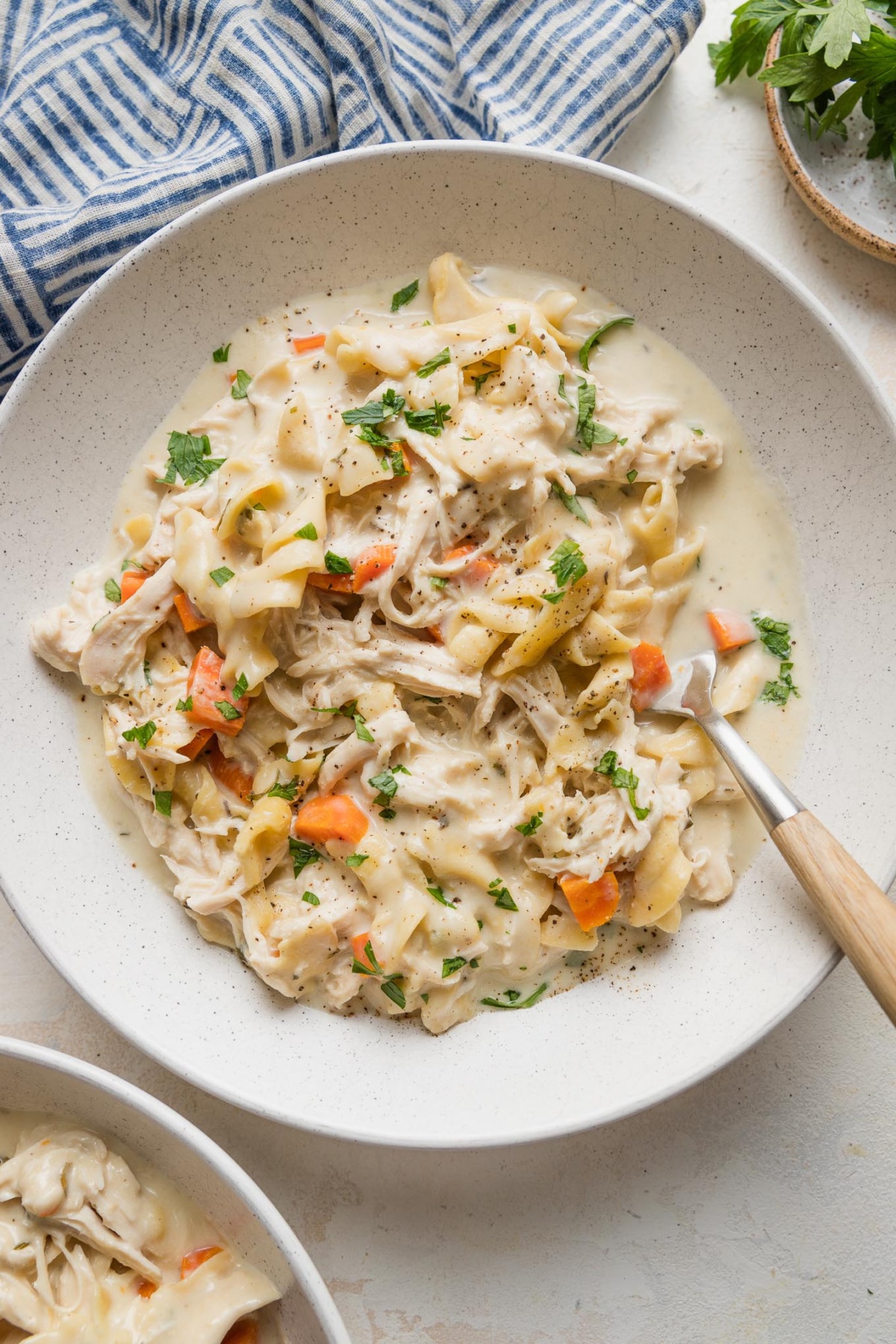 Flat dinner bowl filled with creamy chicken and noodles, with a fork stuck into the helping and a blue napkin on the counter nearby.
