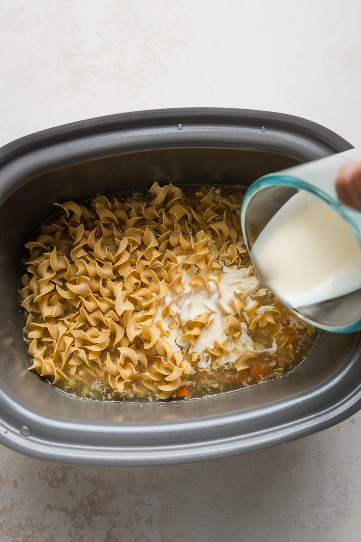 Egg noodles and cream-cornstarch mixture being poured into a slow cooker by a woman's hand.