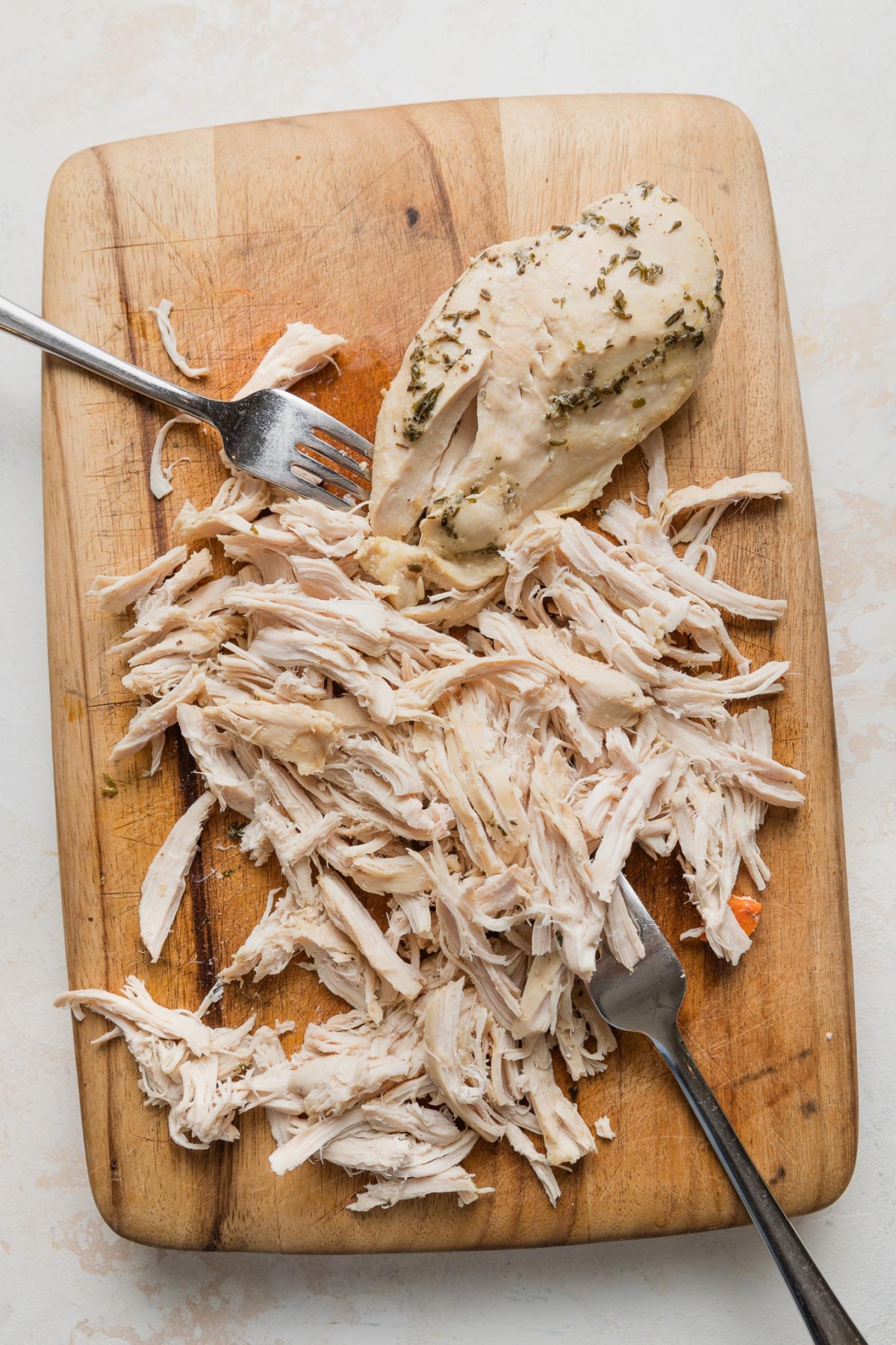 Partially shredded chicken with two forks on a wooden cutting board.
