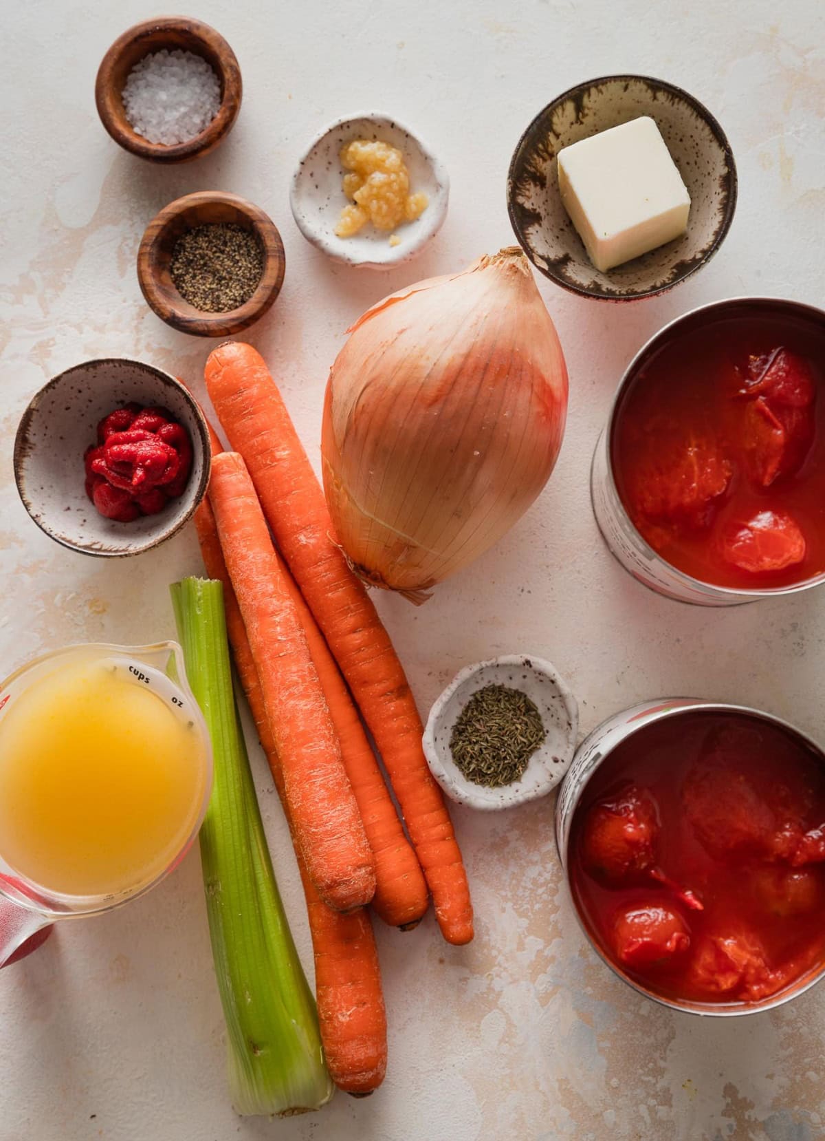Flatlay image of ingredients for a simple homemade tomato soup- canned whole peeled tomatoes, onion, garlic, butter, spices, broth, and carrots and celery to blitz in for good measure.