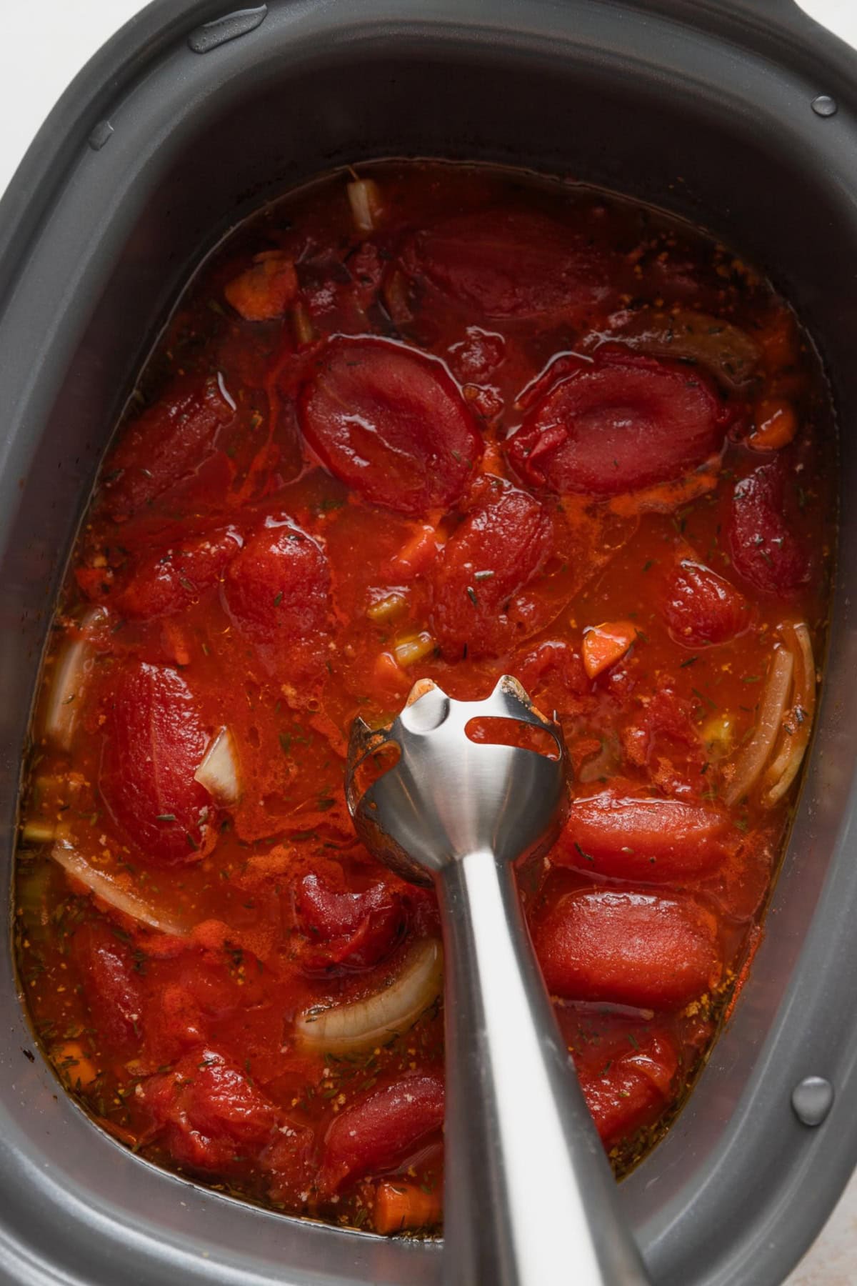 Immersion blender in the midst of pulsing a tomato soup smooth inside of a slow cooker.