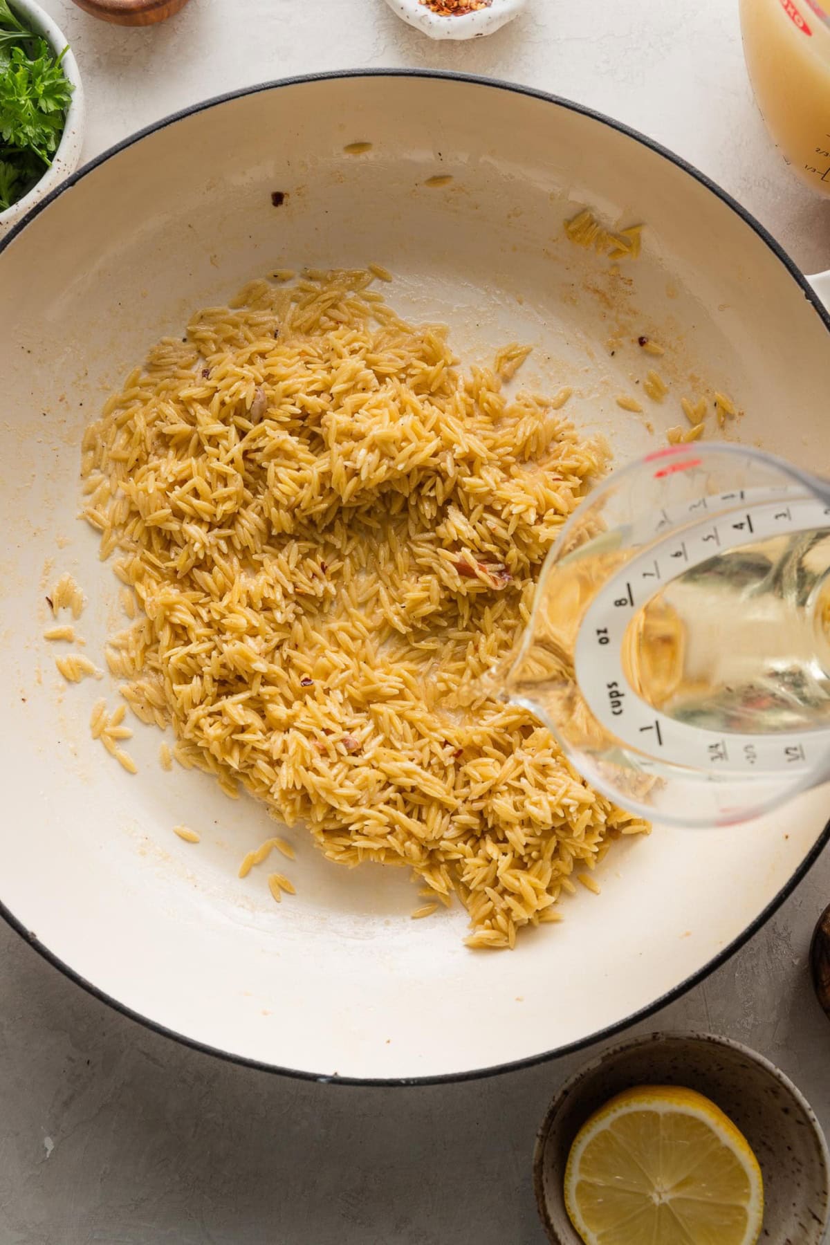 Woman's hand pouring white wine from a liquid measuring cup into the pan of orzo.