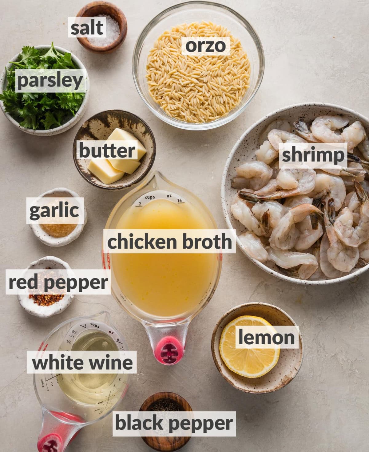 Labeled overhead photo of raw shrimp, uncooked orzo, butter, minced garlic, parsley, chicken broth, white wine, half a lemon, salt, black, and red pepper flakes, all laid on a counter and ready to assemble into a dish of shrimp scampi.