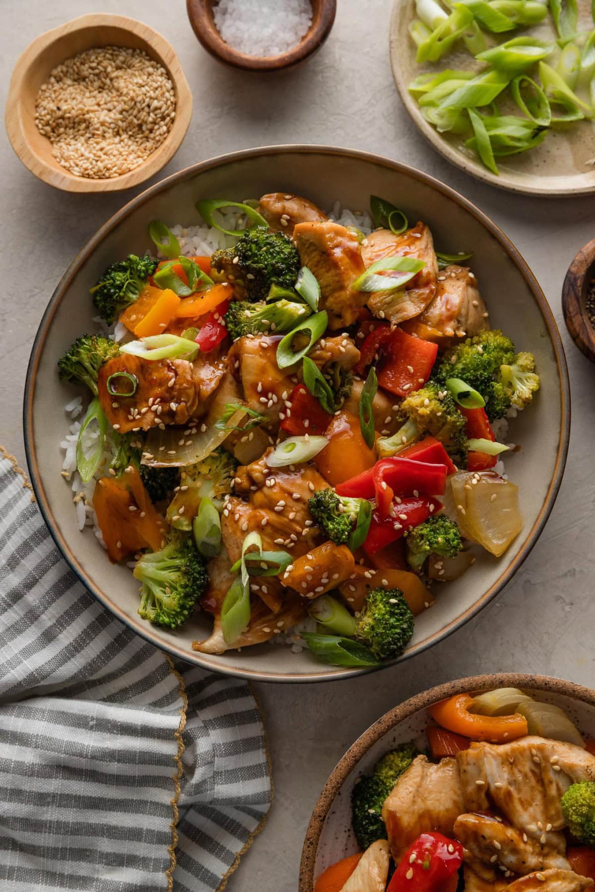Bowl full of sheet pan chicken teriyaki served over rice with sesame seeds and green onions, with a pretty striped napkin and extra garnishes on the counter nearby.