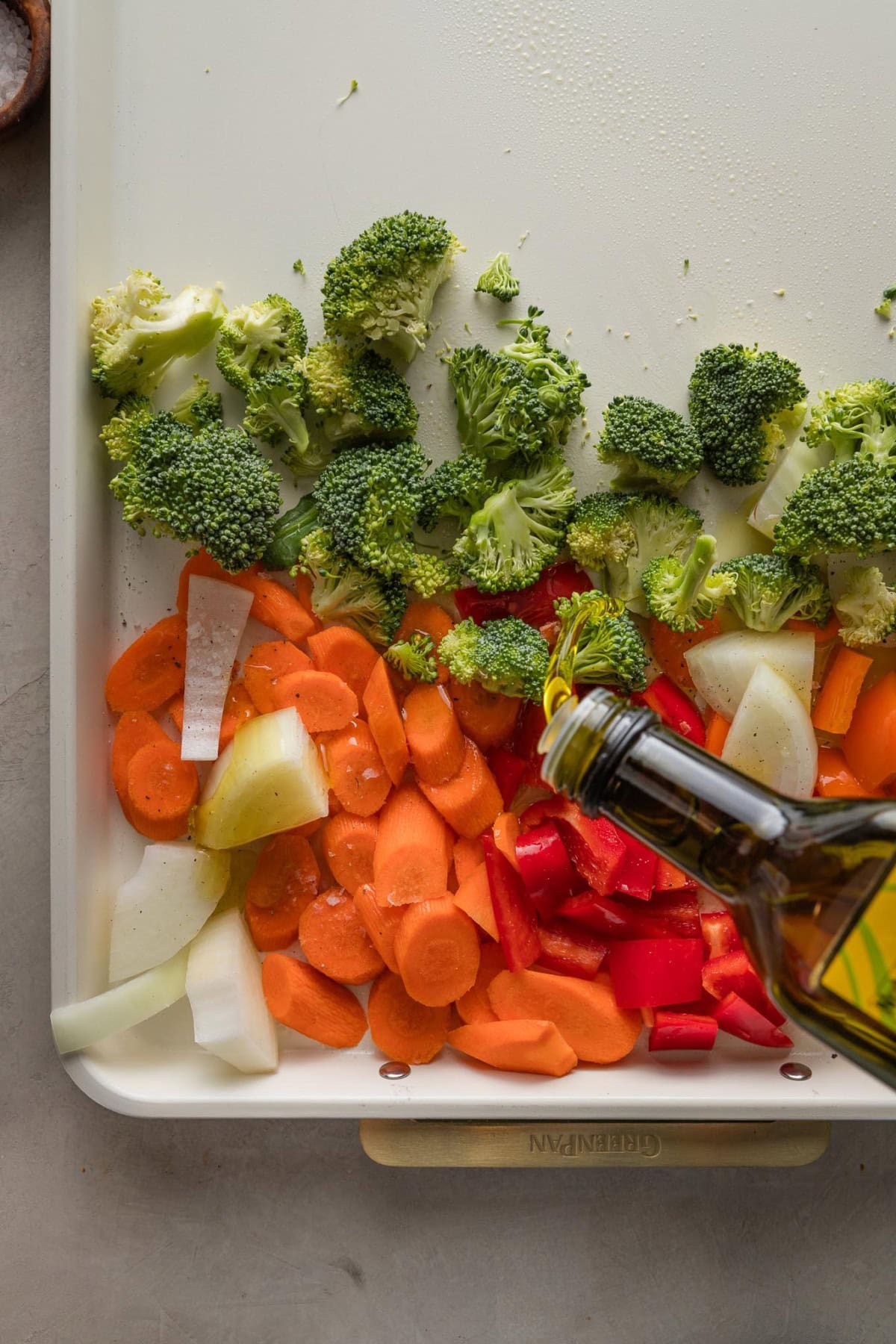Broccoli florets, sliced carrots, bell pepper, and yellow onion tossed onto a large sheet pan, and a woman's hand drizzling olive oil over all the veggies.