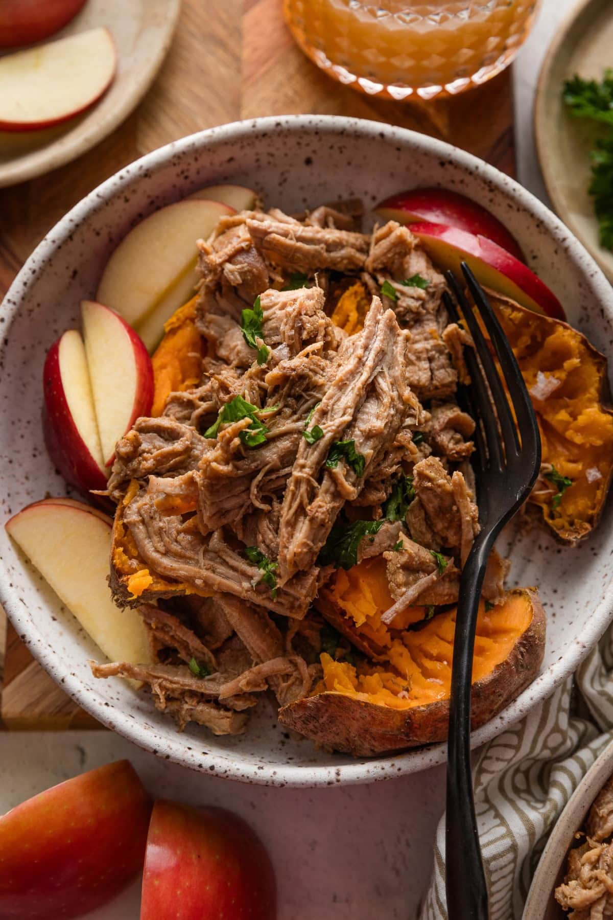 Bowl full of baked sweet potatoes with flaked sea salt, apple slices, and apple cider pulled pork on top with a garnish of fresh parsley.