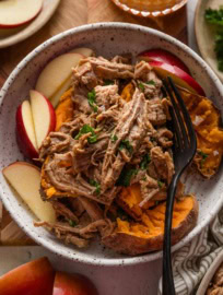 Bowl full of baked sweet potatoes with flaked sea salt, apple slices, and apple cider pulled pork on top with a garnish of fresh parsley.