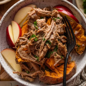 Bowl full of baked sweet potatoes with flaked sea salt, apple slices, and apple cider pulled pork on top with a garnish of fresh parsley.