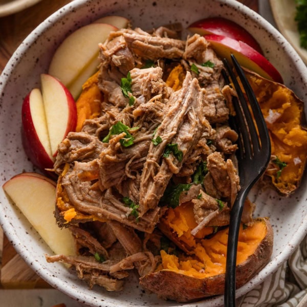 Bowl full of baked sweet potatoes with flaked sea salt, apple slices, and apple cider pulled pork on top with a garnish of fresh parsley.