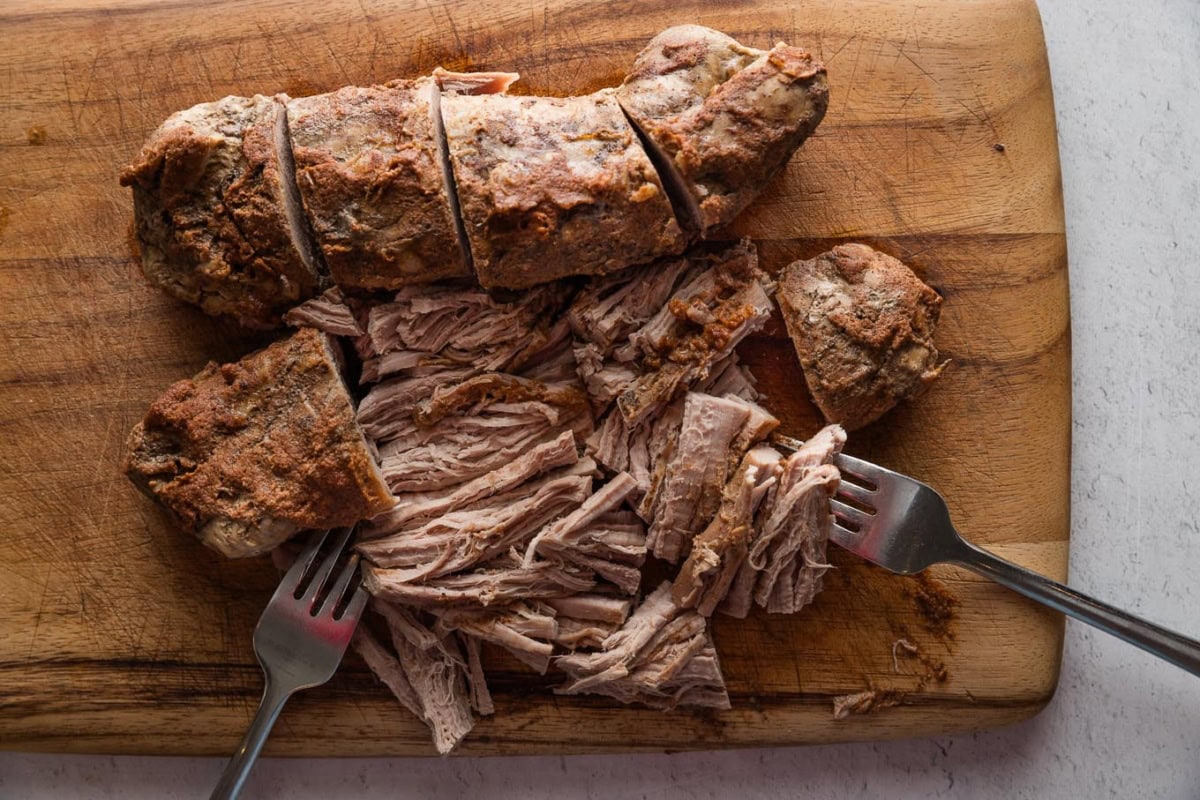 Two pork tenderloins removed from a slow cooker and placed on a cutting board, partly cut and shredded into bite-sized pieces.